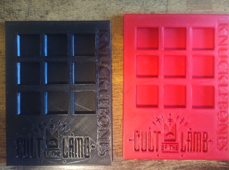 May include: Two 3D printed game trays, one black and one red, with a grid of nine squares. Both trays feature the text "Cult of the Lamb" and "Knick Knack Bones".