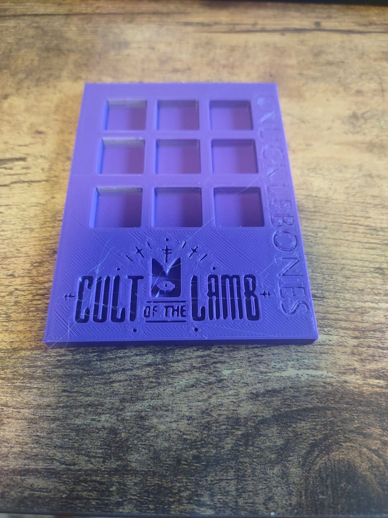 May include: Purple plastic organizer with 9 square compartments. The organizer has the text "Cult of the Lamb" and a logo with a lamb and a cross.