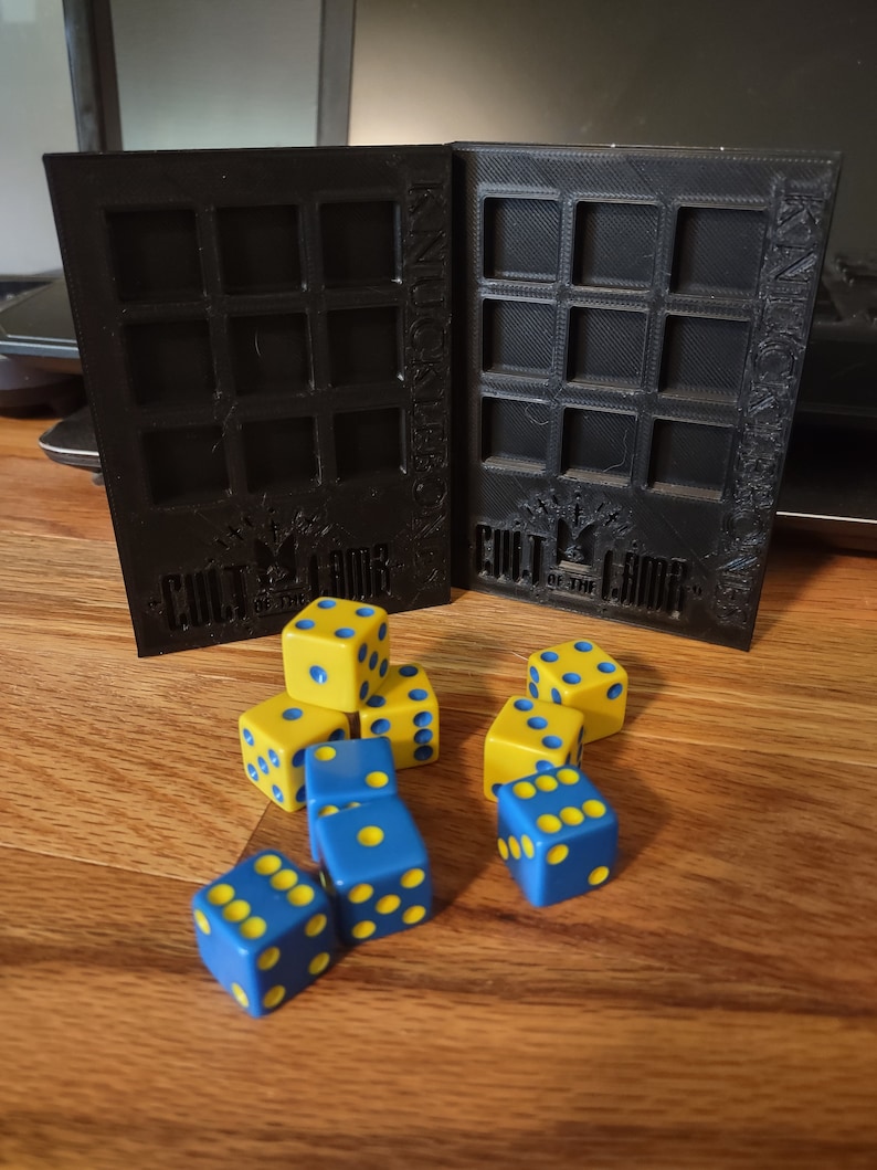 May include: A set of blue and yellow dice with black dots. The dice are on a wooden surface in front of a black 3D printed box with the words "Cult of the Lamb" and "Knucklebones" printed on it.