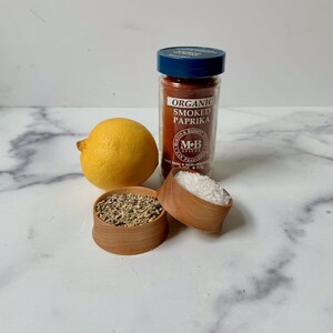 Puede incluir: Un tarro de vidrio de piment&oacute;n ahumado org&aacute;nico con tapa azul, un peque&ntilde;o cuenco de madera lleno de sal, un peque&ntilde;o cuenco de madera lleno de granos de pimienta negra y un lim&oacute;n amarillo sobre una superficie de m&aacute;rmol blanco.