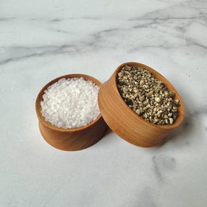 Puede incluir: Dos cuencos de madera llenos de sal blanca y granos de pimienta negra. Los cuencos est&aacute;n sobre una superficie de m&aacute;rmol blanco.