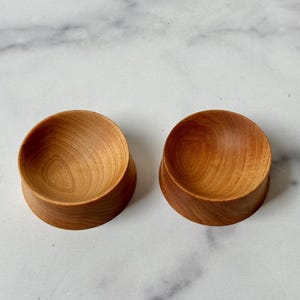 Puede incluir: Dos cuencos de madera peque&ntilde;os, uno ligeramente m&aacute;s grande que el otro, est&aacute;n hechos de madera marr&oacute;n claro con un acabado liso y pulido. Los cuencos son redondos y tienen una ligera curva en sus lados.
