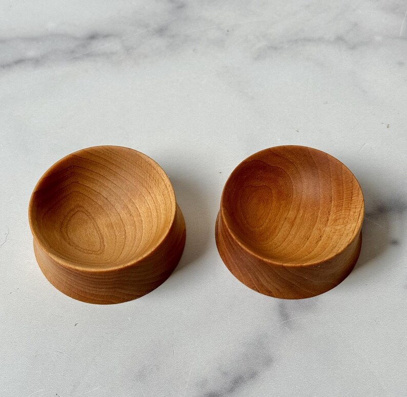Puede incluir: Dos cuencos de madera peque&ntilde;os, uno ligeramente m&aacute;s grande que el otro, est&aacute;n hechos de madera marr&oacute;n claro con un acabado liso y pulido. Los cuencos son redondos y tienen una ligera curva en sus lados.
