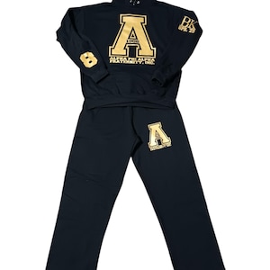 May include: A black hoodie and matching pants set. The hoodie features a large gold "A" with "1906" and "ALPHA PHI ALPHA FRATERNITY, INC." printed below. The pants have a gold "A" on the left leg. The number "8" is on the left sleeve.