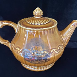 May include: A brown ceramic teapot with a detailed floral design and a winter scene with a house and figures. The teapot has a lid with a knob on top.