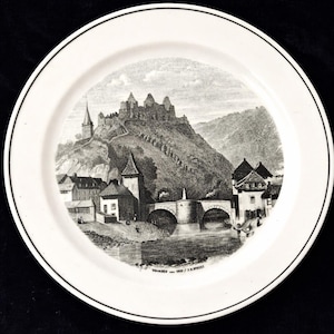 May include: White ceramic plate with a black rim, featuring a black and white illustration of a European town with a castle on a hill, a bridge, and a river. The plate has the text "VANGEN - 1859 / J.B. BREIST" at the bottom.