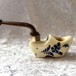 May include: A novelty pipe shaped like a Dutch clog. The pipe is cream-coloured with blue floral designs and a dark brown stem. The metal band around the bowl is silver.