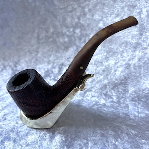 Peut inclure: Une pipe à tabac marron foncé avec une tige incurvée et un support en métal argenté. Le foyer de la pipe est d'un brun profond et riche, et la tige est d'une teinte plus claire. Le support en métal offre une base stable.
