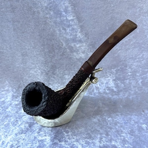 May include: A dark brown tobacco pipe with a textured bowl and a curved, smooth stem. The pipe rests in a silver-coloured metal stand. The bowl is dark, and the stem is a lighter brown colour.