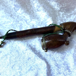 May include: A vintage tobacco pipe with a curved stem and a rounded bowl. The pipe is primarily brown with metal accents and a green string detail. The pipe is resting on a white, textured surface.