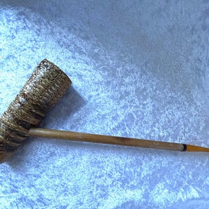 Puede incluir: Una pipa de tabaco artesanal con un cuenco cilíndrico texturizado en tonos marrones y beige. La pipa presenta un largo y delgado vástago de madera con una boquilla curva de color ámbar. El cuenco tiene un patrón único y orgánico.