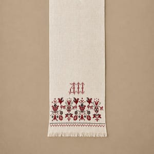Peut inclure: Un chemin de table rectangulaire blanc cassé avec une bordure au point de croix en fil rouge foncé et noir. Le motif comprend des oiseaux stylisés et des fleurs au-dessus d'un bord à franges. Les lettres AM sont brodées en rouge au-dessus.
