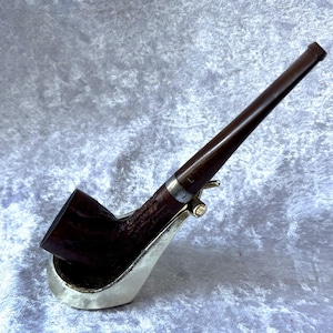 May include: A dark brown tobacco pipe with a long stem, resting on a silver-coloured stand. The pipe has a curved bowl and a metal band near the stem's connection. The stand is shaped to cradle the pipe.