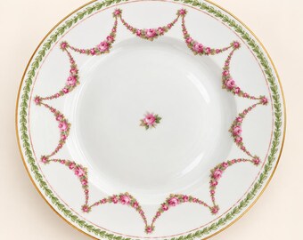 May include: A white porcelain dinner plate featuring a gold rim, a green leaf garland border, and a series of pink rose floral swags draped around the rim. A single pink rose blossom is centered in the middle of the plate.
