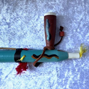 May include: A decorative wooden pipe with a turquoise body, decorated with black, brown, and yellow accents. The pipe features a brown bowl with a black and blue design, a leather strap, and red and yellow feathers. The mouthpiece is white.
