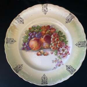 Puede incluir: Un plato de porcelana vintage con un esmalte verde y blanco. El plato presenta un diseño pintado a mano de fruta, incluyendo uvas, melocotones y cerezas. El plato está decorado con detalles en negro y dorado.
