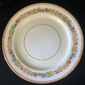 May include: A white porcelain dinner plate with a gold rim and a floral design in shades of pink, yellow, blue, and green.