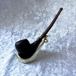 Peut inclure: Une pipe à tabac noire avec un tuyau brun foncé, posée dans un support argenté. La pipe a un design incurvé et est faite d'un matériau foncé et texturé. Le tuyau est long et fin.