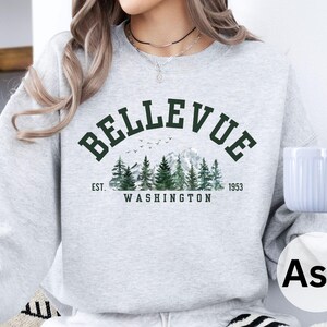 May include: Ash-colored sweatshirt with the word "Bellevue" in dark green arched across the top. Below the word is a graphic of evergreen trees, mountains, and birds. The text "EST. 1953 WASHINGTON" is below the graphic.