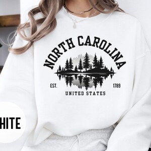 May include: White sweatshirt with a black graphic featuring the words "NORTH CAROLINA" arched above a silhouette of trees and water. Below the graphic are the words "EST. 1789 UNITED STATES". The word "WHITE" is printed in a white circle.