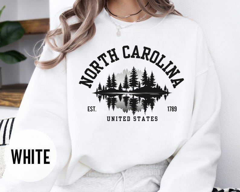 May include: White sweatshirt with a black graphic featuring the words "NORTH CAROLINA" arched above a silhouette of trees and water. Below the graphic are the words "EST. 1789 UNITED STATES". The word "WHITE" is printed in a white circle.