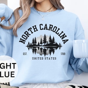 May include: Light blue sweatshirt featuring a black graphic of a forest scene reflected in water, with the text "NORTH CAROLINA" above and "EST. 1789 UNITED STATES" below. The sweatshirt is a solid light blue color.