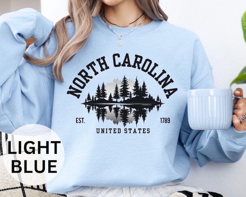 May include: Light blue sweatshirt featuring a black graphic of a forest scene reflected in water, with the text "NORTH CAROLINA" above and "EST. 1789 UNITED STATES" below. The sweatshirt is a solid light blue color.