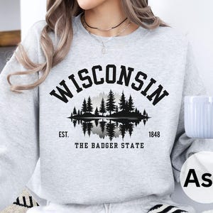 May include: Ash-colored sweatshirt with the word "WISCONSIN" arched across the top in black block letters. Below the word is a black and white graphic of a forest reflected in water. The text "EST. 1848 THE BADGER STATE" is below the graphic.