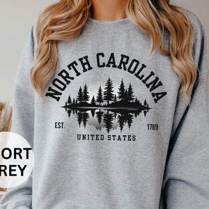May include: Sport grey sweatshirt featuring a black graphic of a North Carolina landscape with trees and a lake, with the text "NORTH CAROLINA" arched above the image. Below the image are the words "EST. UNITED STATES 1789".
