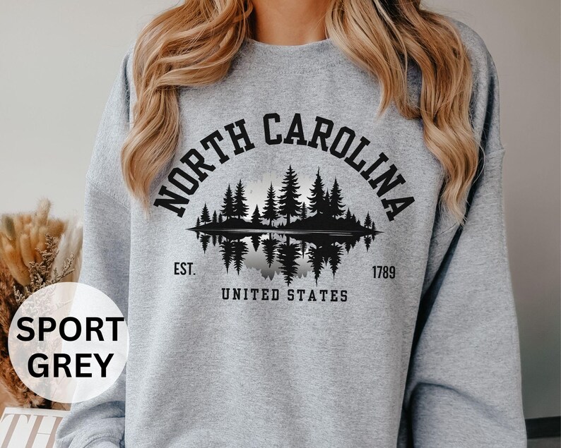 May include: Sport grey sweatshirt featuring a black graphic of a North Carolina landscape with trees and a lake, with the text "NORTH CAROLINA" arched above the image. Below the image are the words "EST. UNITED STATES 1789".
