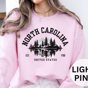 May include: Light pink sweatshirt with a black graphic of a forest scene and the words "NORTH CAROLINA" in an arch. Below the graphic are the words "EST. 1789 UNITED STATES". The sweatshirt is a solid light pink color.