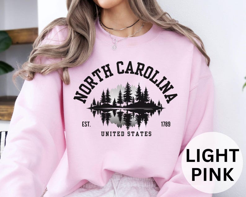 May include: Light pink sweatshirt with a black graphic of a forest scene and the words "NORTH CAROLINA" in an arch. Below the graphic are the words "EST. 1789 UNITED STATES". The sweatshirt is a solid light pink color.