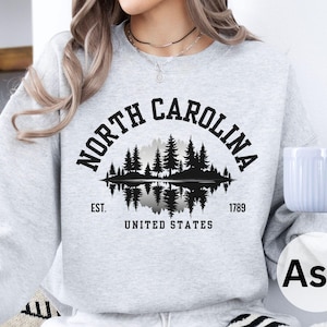 May include: Ash gray sweatshirt with a black graphic of a North Carolina landscape, including trees and a lake, with the text "NORTH CAROLINA" above and "UNITED STATES" below. The sweatshirt also has the text "EST. 1789".