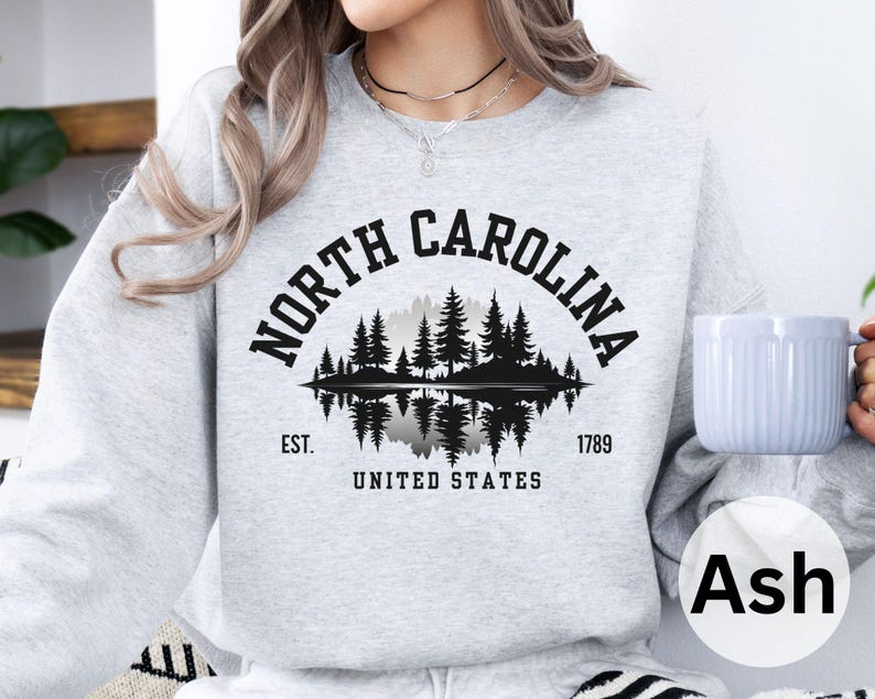 May include: Ash gray sweatshirt with a black graphic of a North Carolina landscape, including trees and a lake, with the text "NORTH CAROLINA" above and "UNITED STATES" below. The sweatshirt also has the text "EST. 1789".