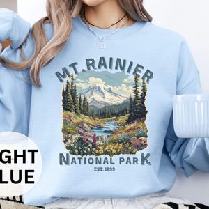 May include: Light blue sweatshirt with a graphic of Mount Rainier National Park, featuring a mountain scene with trees and a river. The text "MT. RAINIER NATIONAL PARK EST. 1899" is printed on the sweatshirt.