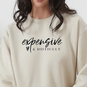 May include: A beige sweatshirt with the text "expensive & difficult" in black script with a small heart symbol.