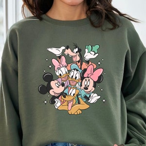 K&ouml;nnte beinhalten: Gr&uuml;ner Pullover mit einem Aufdruck von Mickey Mouse, Minnie Mouse, Donald Duck, Goofy und Pluto. Die Figuren l&auml;cheln alle und scheinen sich zu umarmen.