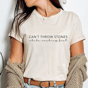 May include: A beige t-shirt with the text "CAN'T THROW STONES while washing feet" printed on the front.