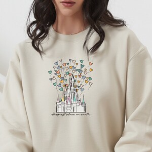 May include: A beige sweatshirt with a graphic of Cinderella's castle and colorful Mickey Mouse balloons. The text "Happiest place on earth" is printed below the castle.
