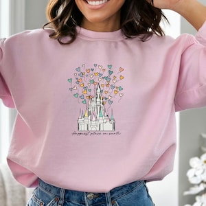 May include: A pink sweatshirt with a graphic of Cinderella's castle and colorful balloons shaped like Mickey Mouse heads. The text "Happiest place on earth" is printed below the castle.