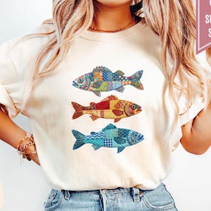 May include: An ivory t-shirt featuring a graphic print of three fish stacked vertically. Each fish displays a different colorful patchwork pattern with various geometric designs. A pink banner in the top right corner contains the text SUPER SOFT.