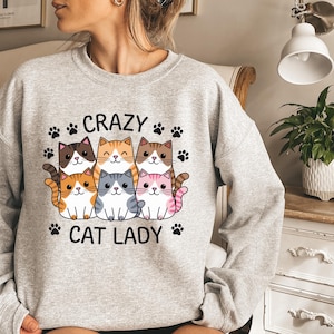 May include: A light gray sweatshirt featuring a graphic of six cartoon cats in various colors. The text "CRAZY CAT LADY" is printed above and below the cats, with paw prints scattered around the text. The sweatshirt is made of a soft material.