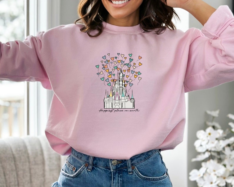 May include: A pink sweatshirt with a graphic of Cinderella's castle and colorful balloons shaped like Mickey Mouse heads. The text "Happiest place on earth" is printed below the castle.