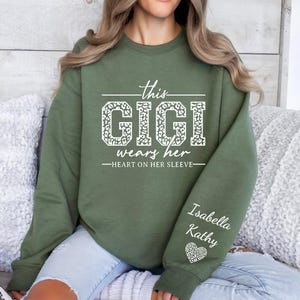 May include: Green sweatshirt with the text "This Gigi wears her heart on her sleeve" in white with a leopard print design. The sleeve has the names "Isabella Kathy" and a heart with a leopard print design.