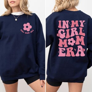 Puede incluir: Sudadera azul marino con el texto "GIRL MOM ERA" y un gr&aacute;fico de flores rosas en la parte delantera. La parte trasera de la sudadera presenta el texto "IN MY GIRL MOM ERA" en rosa, con letras de estilo retro. Una prenda superior informal y c&oacute;moda.