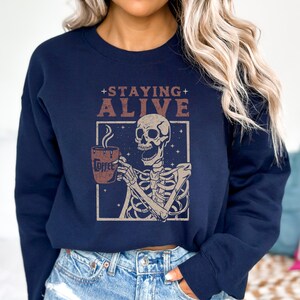 K&ouml;nnte beinhalten: Marineblaues Sweatshirt mit einem Skelett, das eine Kaffeetasse h&auml;lt. Der Aufdruck enth&auml;lt die Worte "Staying Alive" und "Coffee". Dieses l&auml;ssige Sweatshirt ist ein bequemes Kleidungsst&uuml;ck, das sich f&uuml;r den Alltag eignet.