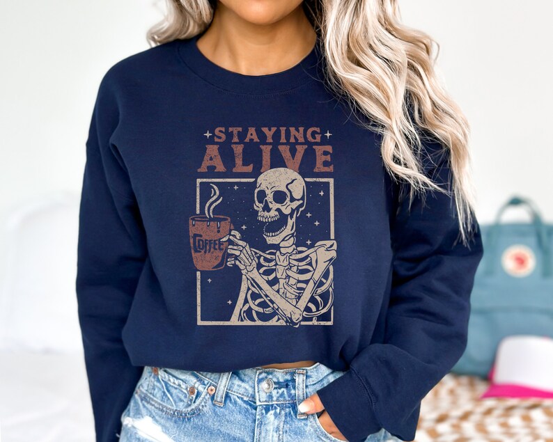K&ouml;nnte beinhalten: Marineblaues Sweatshirt mit einem Skelett, das eine Kaffeetasse h&auml;lt. Der Aufdruck enth&auml;lt die Worte "Staying Alive" und "Coffee". Dieses l&auml;ssige Sweatshirt ist ein bequemes Kleidungsst&uuml;ck, das sich f&uuml;r den Alltag eignet.