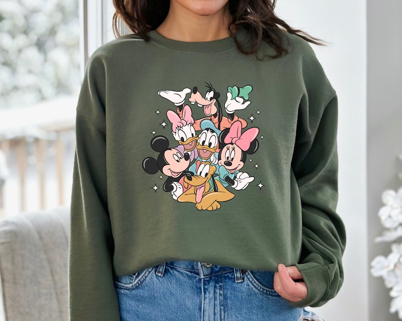K&ouml;nnte beinhalten: Gr&uuml;ner Pullover mit einem Aufdruck von Mickey Mouse, Minnie Mouse, Donald Duck, Goofy und Pluto. Die Figuren l&auml;cheln alle und scheinen sich zu umarmen.