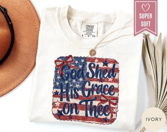 Può includere: Una t-shirt a maniche corte color avorio con una grafica della bandiera americana con bordo smerlato, due fiocchi rossi e il testo blu "God Shed His Grace on Thee". La maglietta ha un'etichetta Comfort Colors sul collo e una collana dorata.