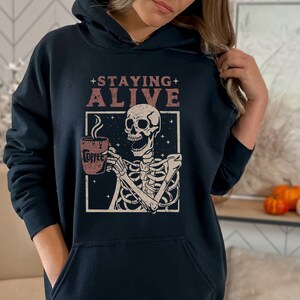 K&ouml;nnte beinhalten: Marineblauer Kapuzenpullover mit einer Grafik eines Skeletts, das eine Kaffeetasse h&auml;lt. Der Text "+ STAYING ALIVE" steht &uuml;ber dem Skelett. Auf der Kaffeetasse steht das Wort "Coffee". Das Design befindet sich in einem wei&szlig;en Rahmen mit Sternenhintergrund.